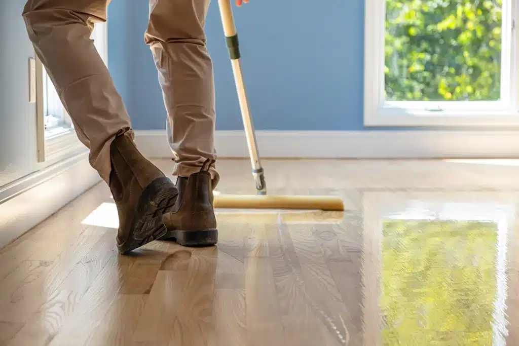 Zoomed in image of a ReCoat Revolution employee's legs from knee down using a foam applicator to spread new finish on a light, hardwood floor.